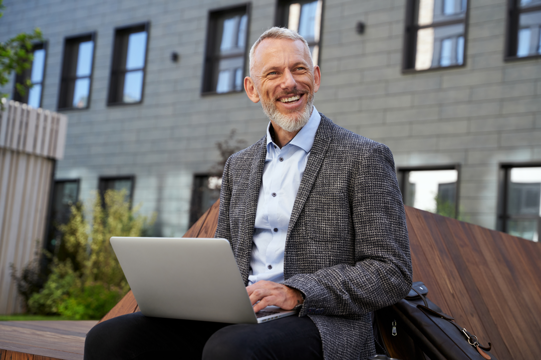 Happy upper middle age business man sitting outside using laptop looking at the camera